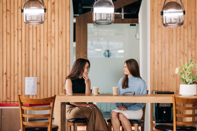 Two women sitting at a café table, smiling and talking closely over coffee