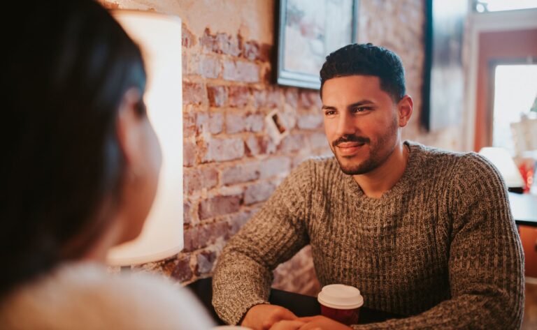Young couple sitting across from each other at a café table, leaning in for a warm, unguarded conversation over coffee