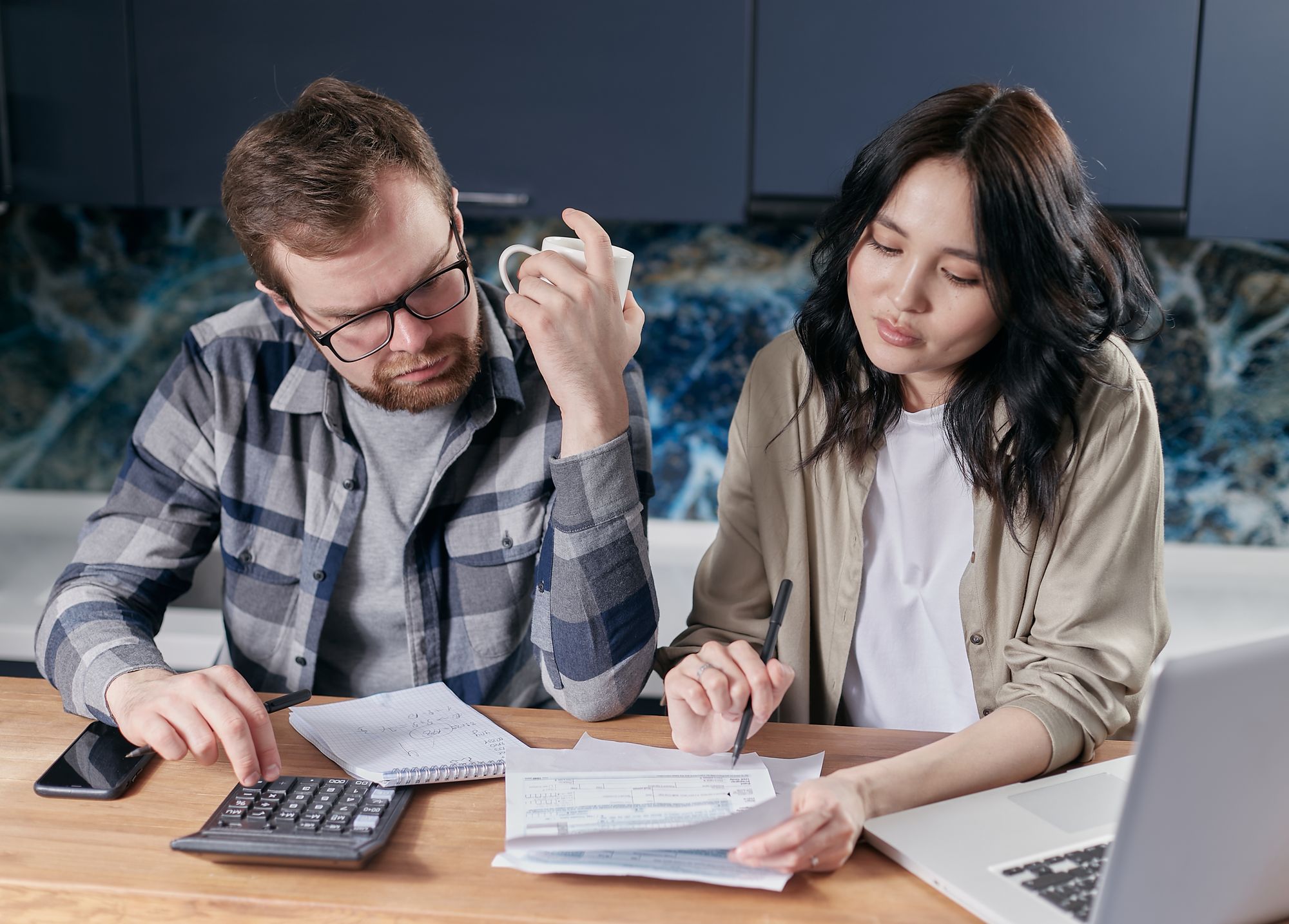 A couple sitting together at a kitchen table in natural daylight, reviewing bills and using a calculator and laptop to go over their household budget