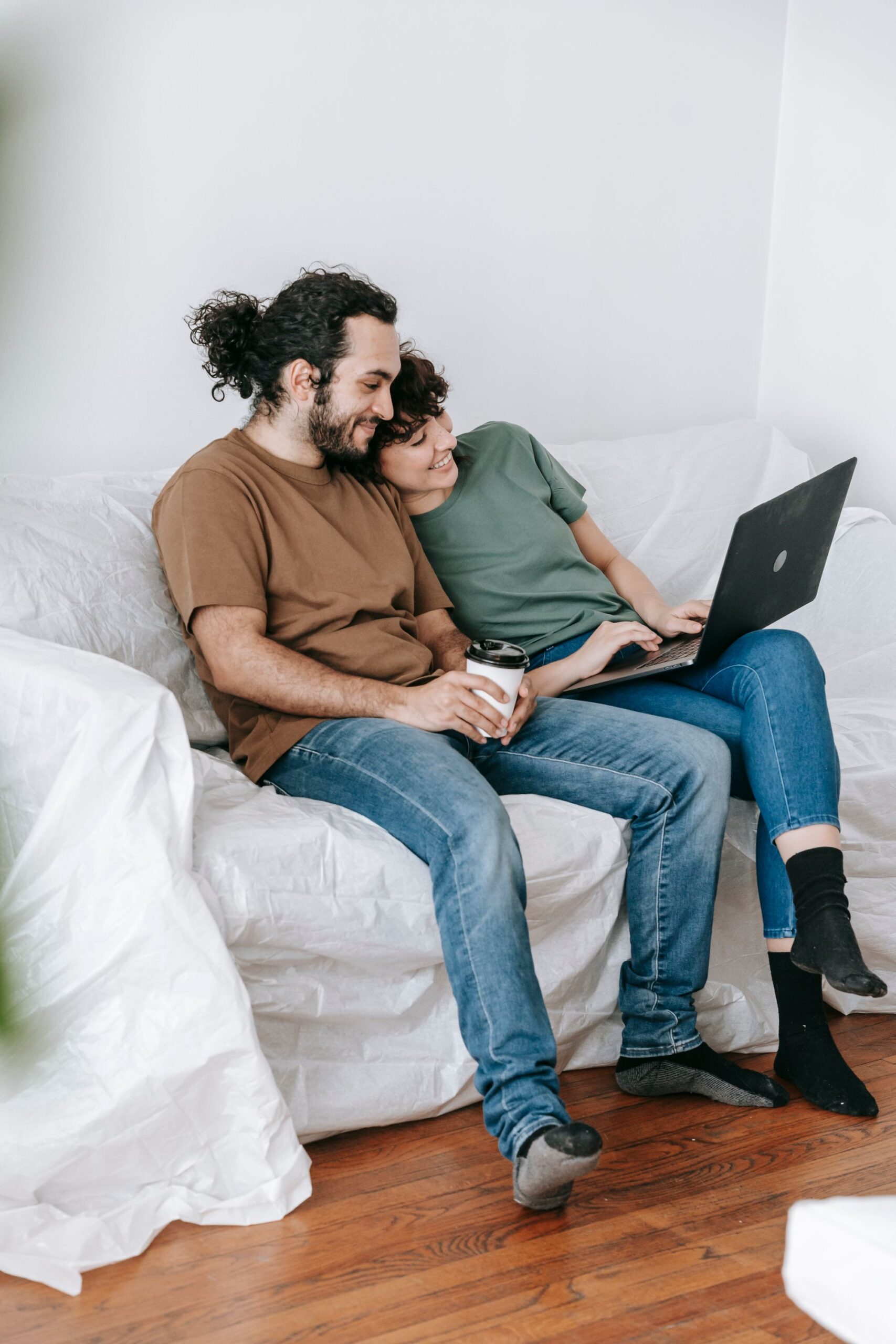 A young couple sitting together on a couch at home, relaxed and casually dressed, looking at a laptop screen together