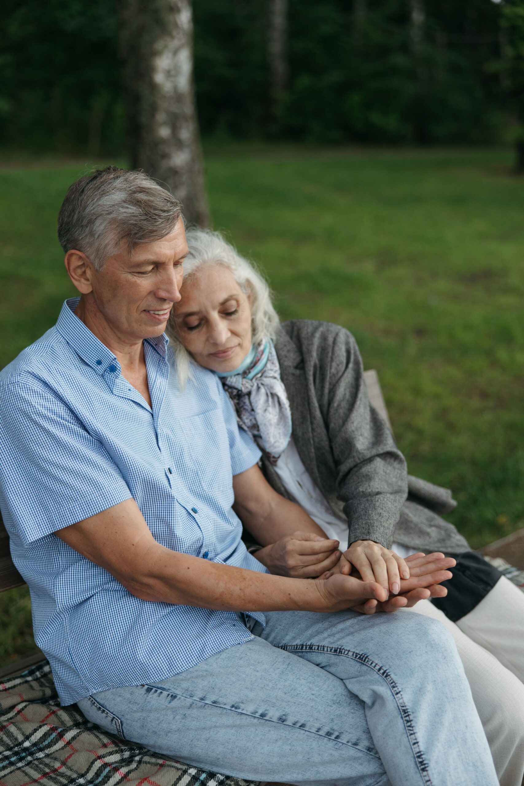 An elderly couple sitting together outdoors on a park bench in soft natural light, surrounded by greenery