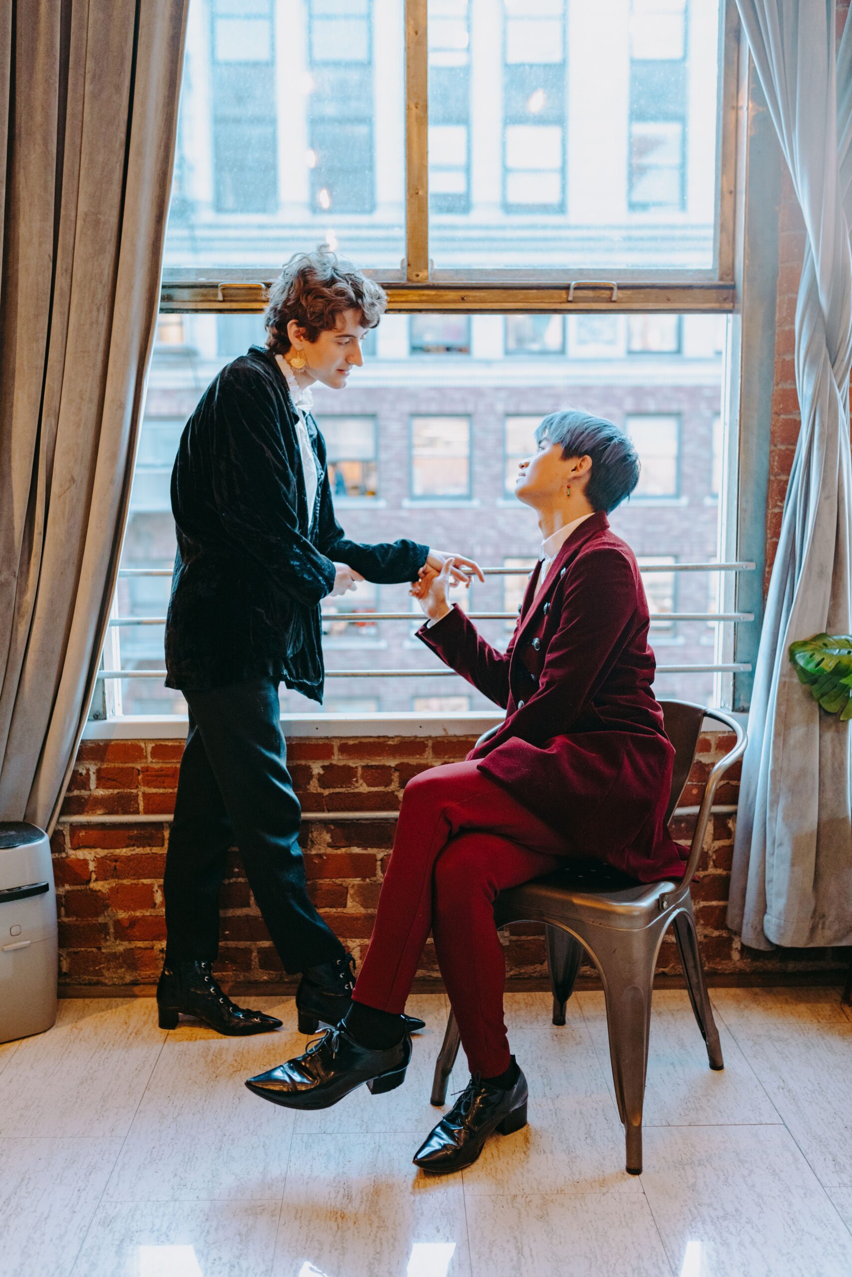 Two men share a tender, sunlit moment together indoors near a window, holding hands and standing close
