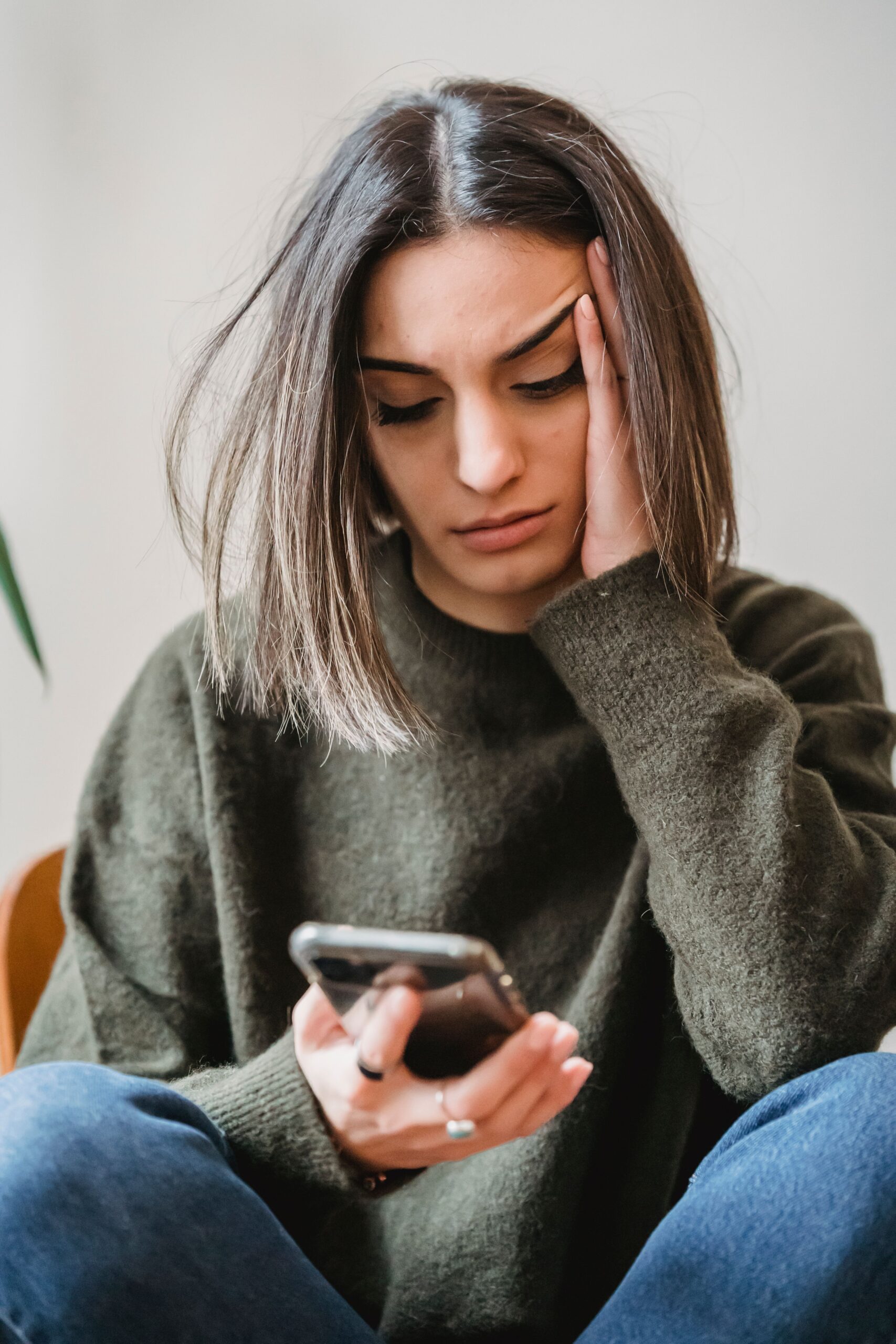 A woman indoors resting her hand against her face with a concerned expression while holding her phone