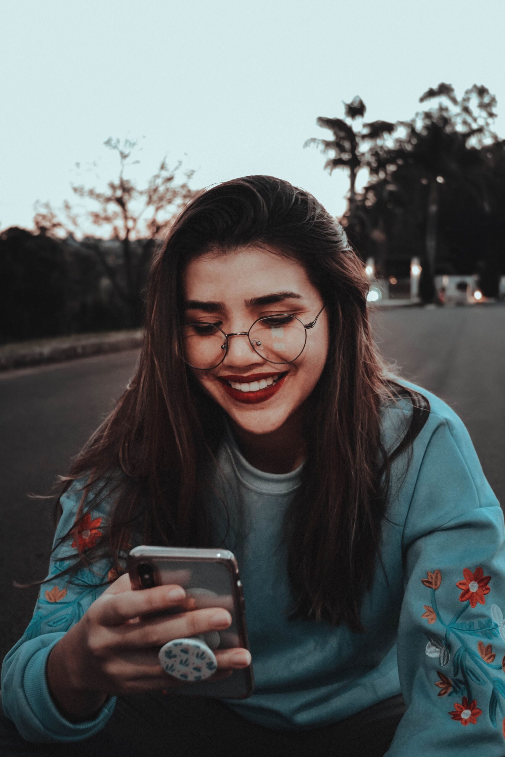 A young woman sitting outdoors, smiling as she looks at her smartphone screen