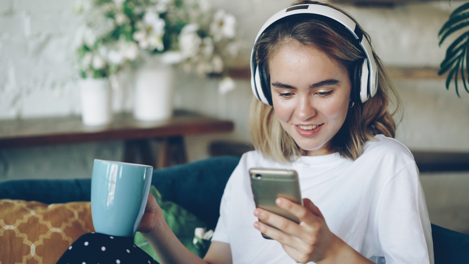 Young woman with headphones holds mug and phone.