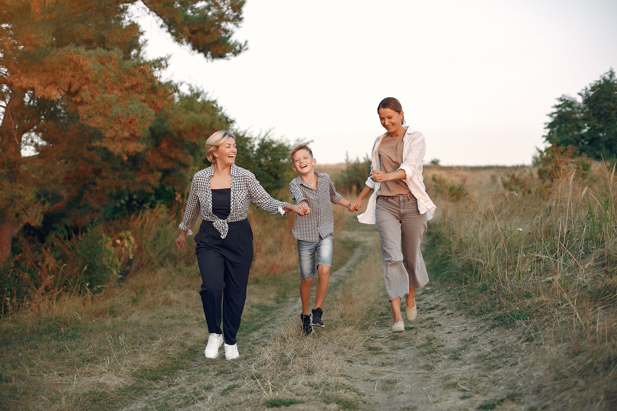 Three generations - a mother, her preteen son, and grandmother - walking hand in hand along a sunlit countryside path, representing sandwich generation caregivers