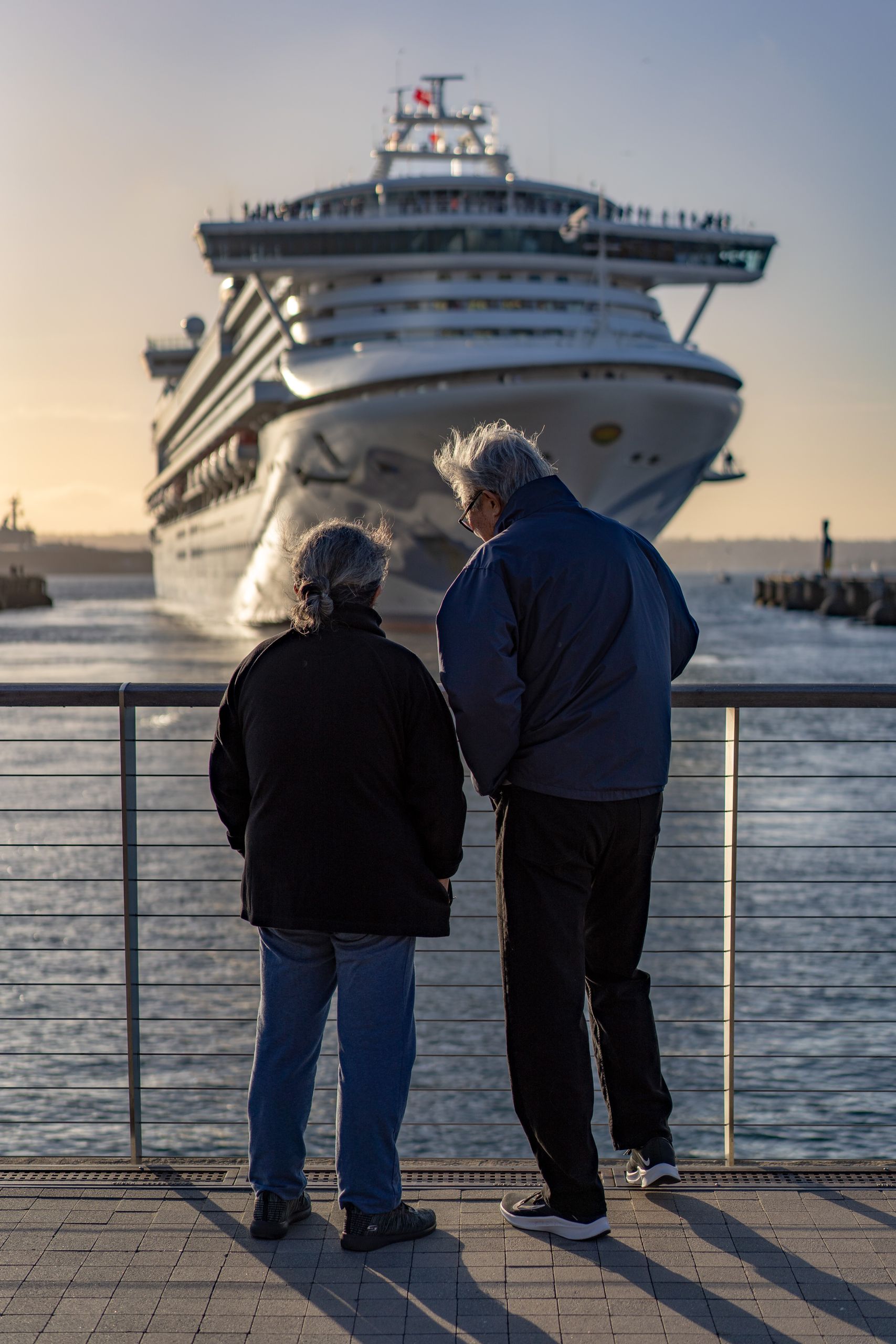 Senior couple standing at railing, admiring large cruise ship approaching dock at sunset.