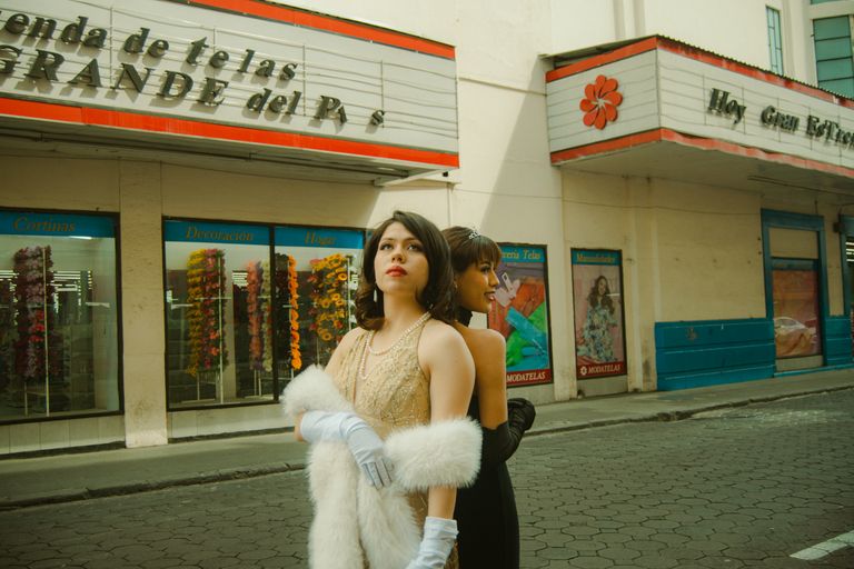 Two elegant women in vintage attire pose outside a textile store, evoking a classic, retro vibe.
