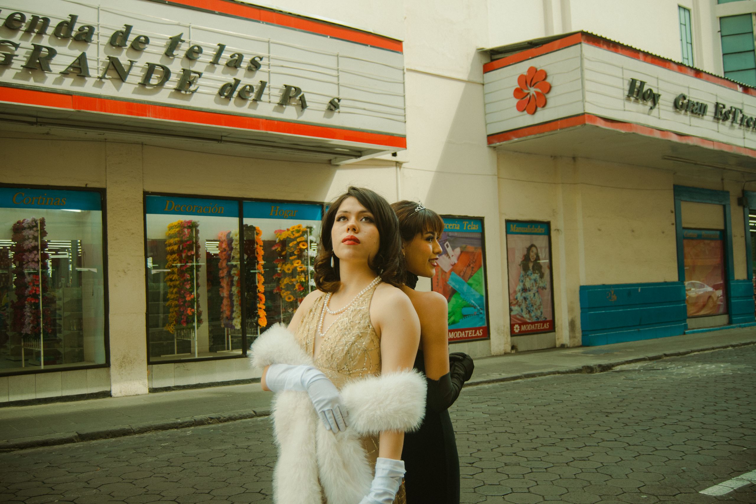 Two elegant women in vintage attire pose outside a textile store, evoking a classic, retro vibe.