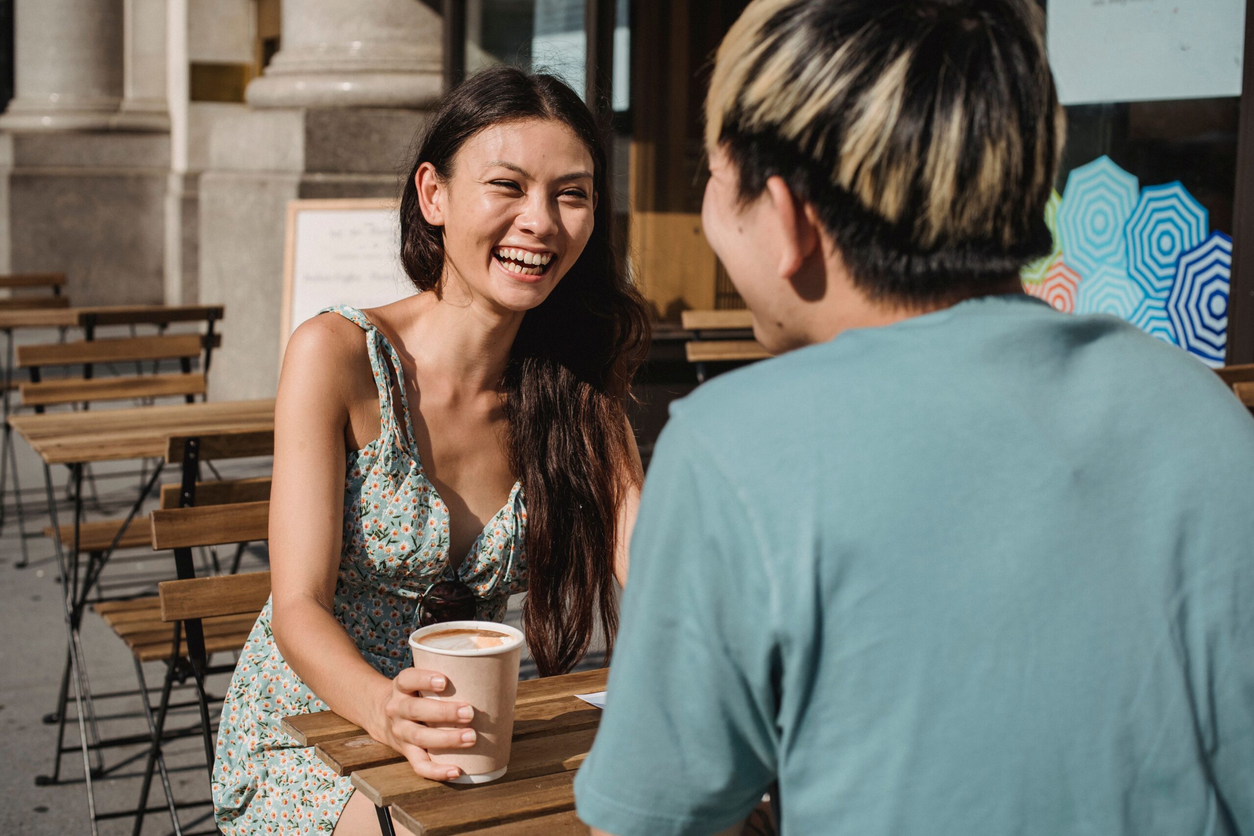 Two friends sitting together at an outdoor cafe table, talking and laughing over coffee in the sun