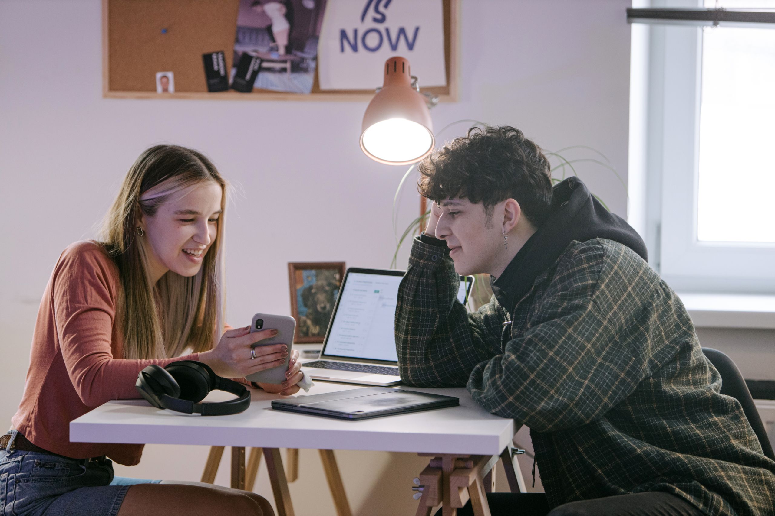 Two young adults engage in conversation, using technology at an indoor desk.
