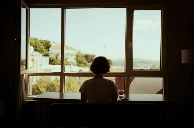 A woman sits alone at a table, gazing out a window in quiet solitude