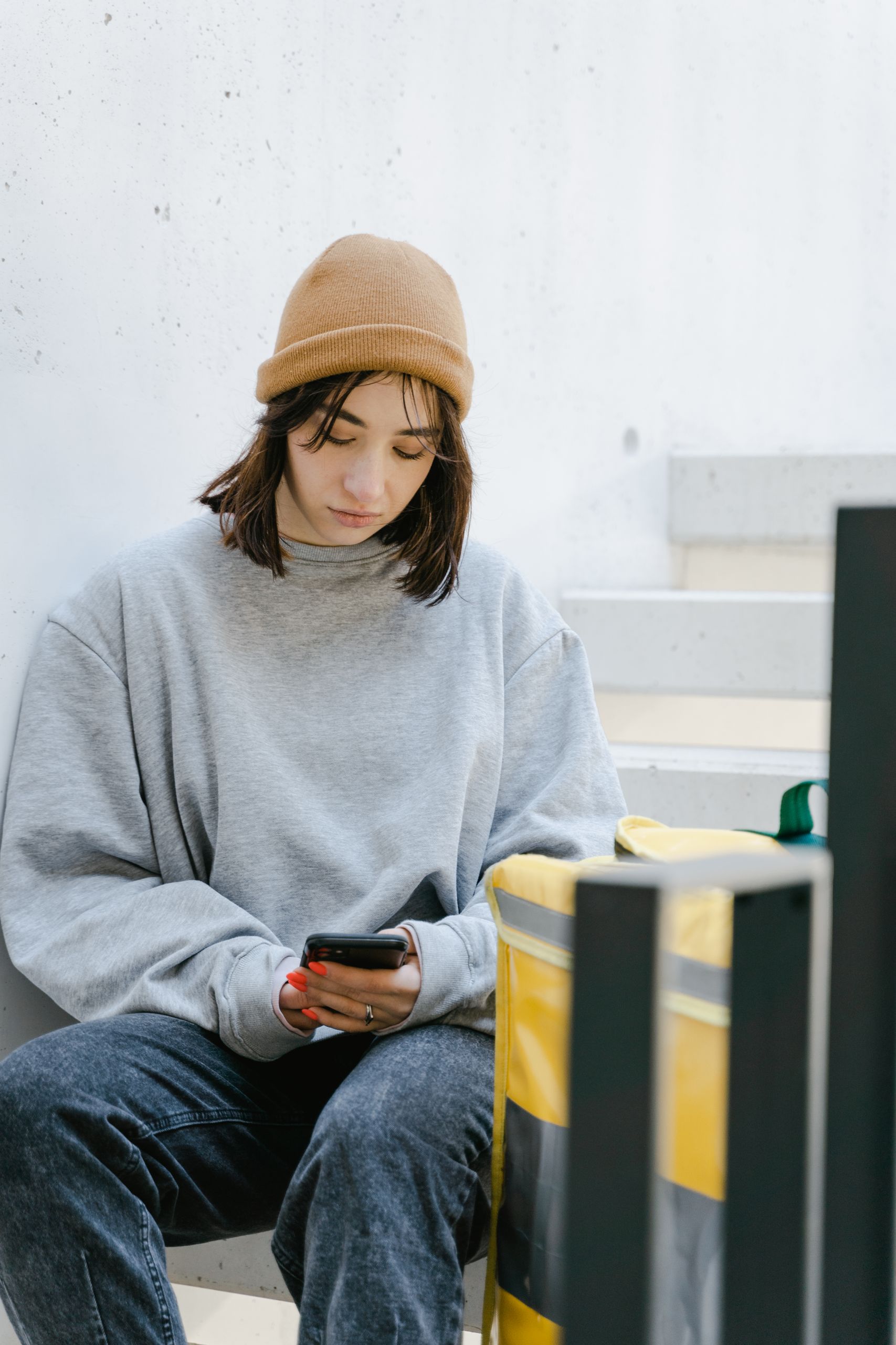Young woman dressed casually with a beanie, checking her phone next to a food delivery bag outdoors.