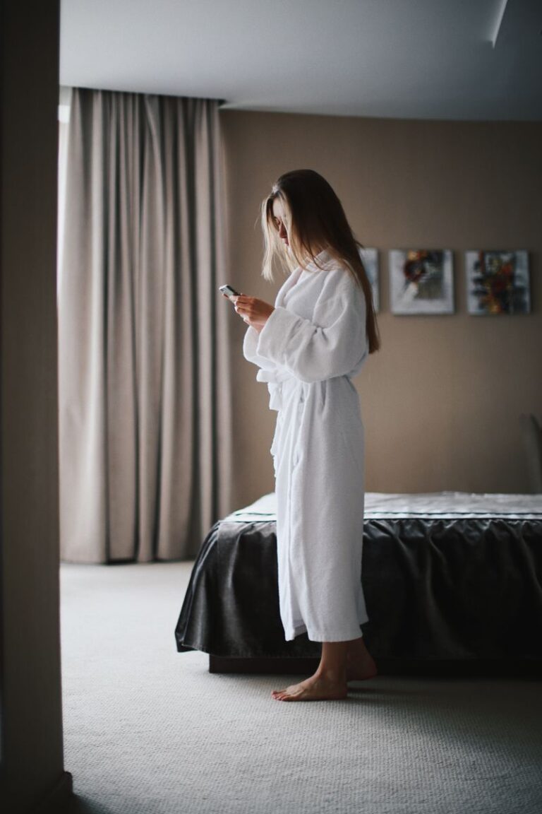Woman standing in a hotel room wearing a bathrobe, focused on her smartphone for communication or relaxation.