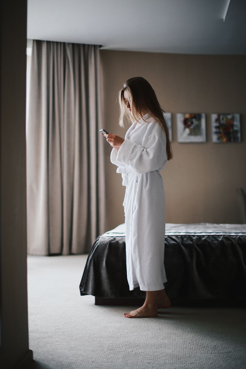 Woman standing in a hotel room wearing a bathrobe, focused on her smartphone for communication or relaxation.