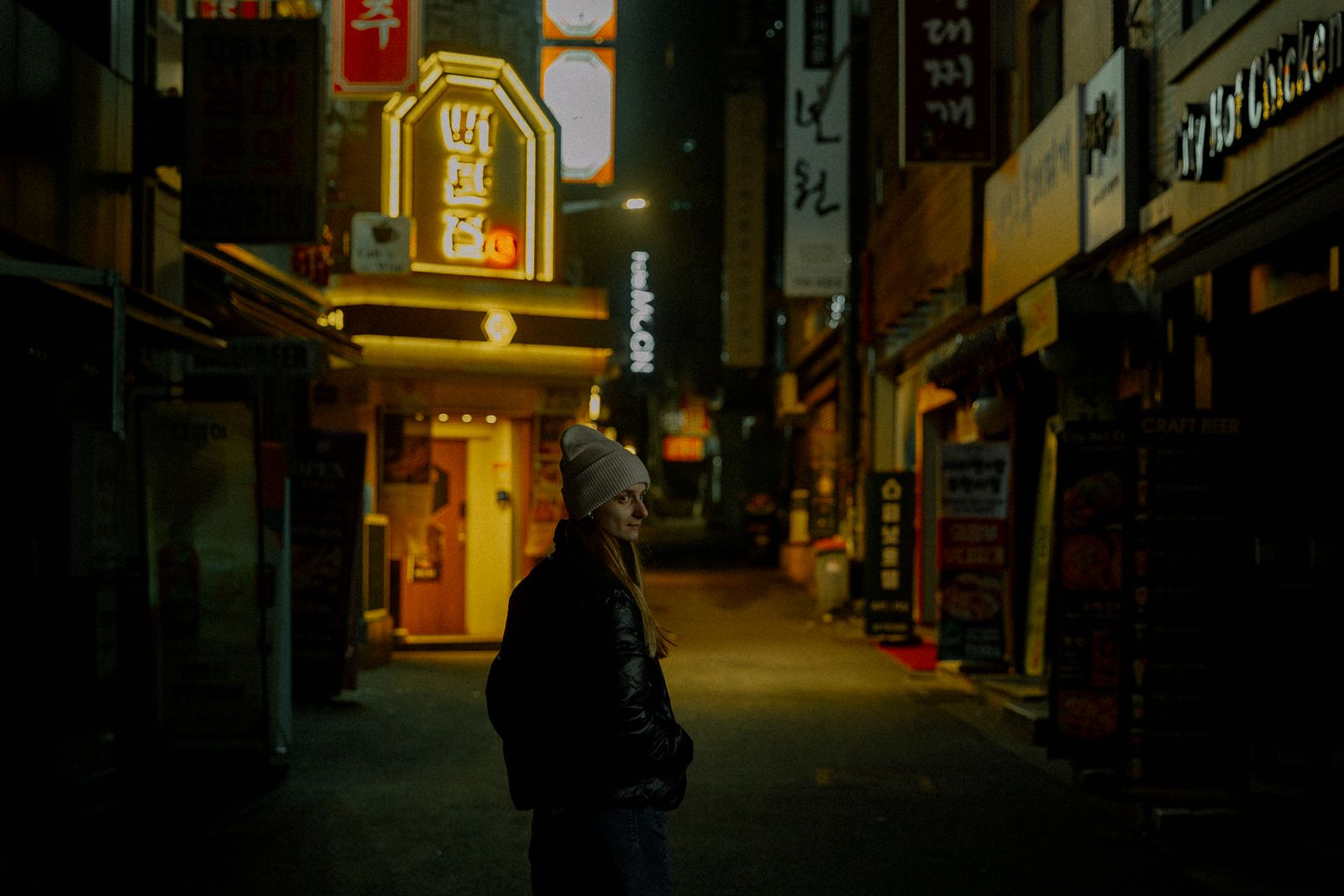 A woman in a beanie and jacket stands in a dimly lit urban alley at night, surrounded by neon signs.