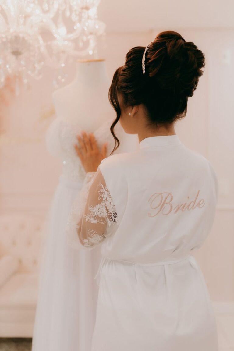 Bride in a white robe admiring wedding gown. Bridal preparation moments.