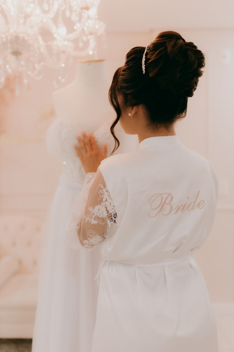 Bride in a white robe admiring wedding gown. Bridal preparation moments.