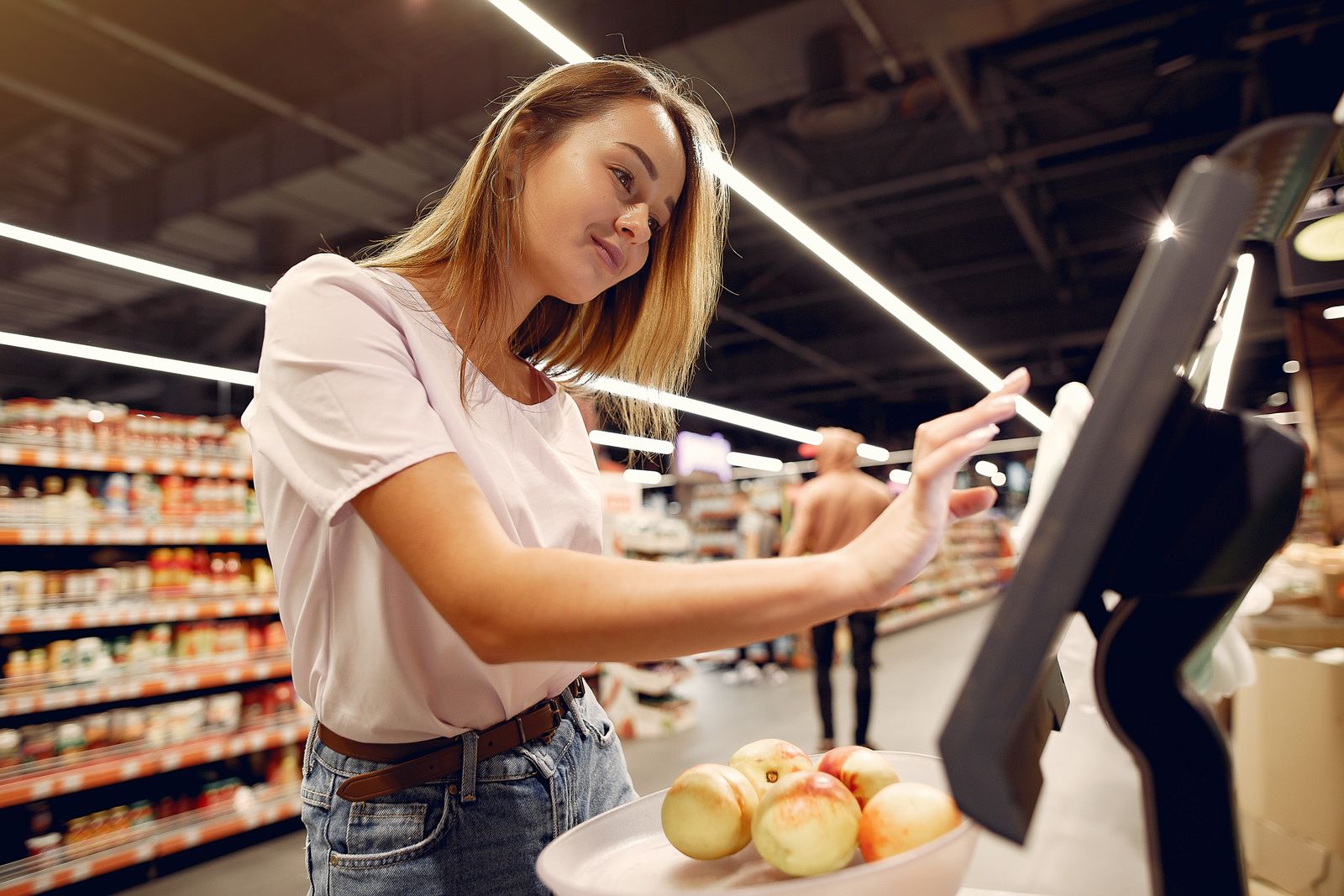 Side view of young woman in trendy clothes weighing peaches on scales while shopping in supermarket during purchase food