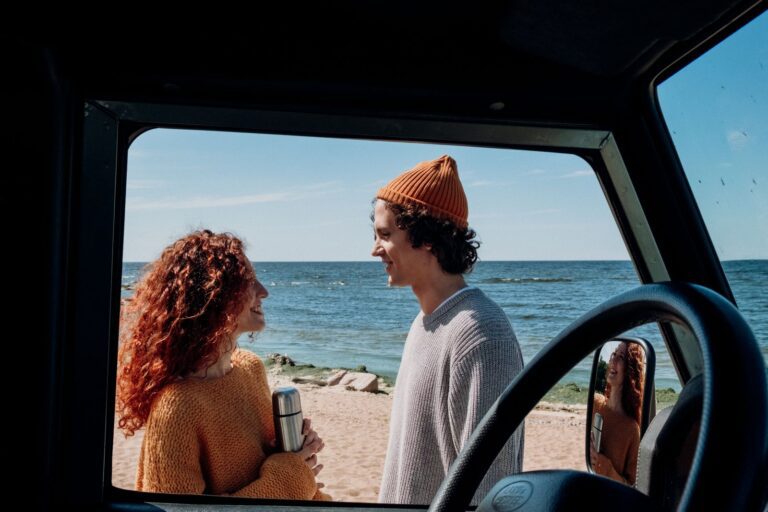 A cheerful couple smiles as they enjoy a sunny day on the beach, framed by a car window.