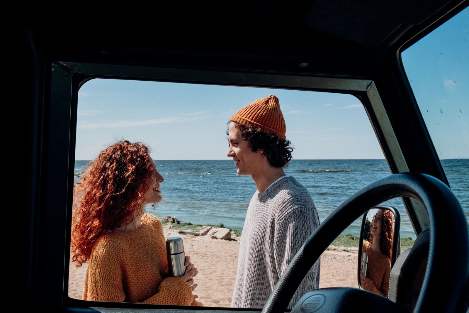 A cheerful couple smiles as they enjoy a sunny day on the beach, framed by a car window.