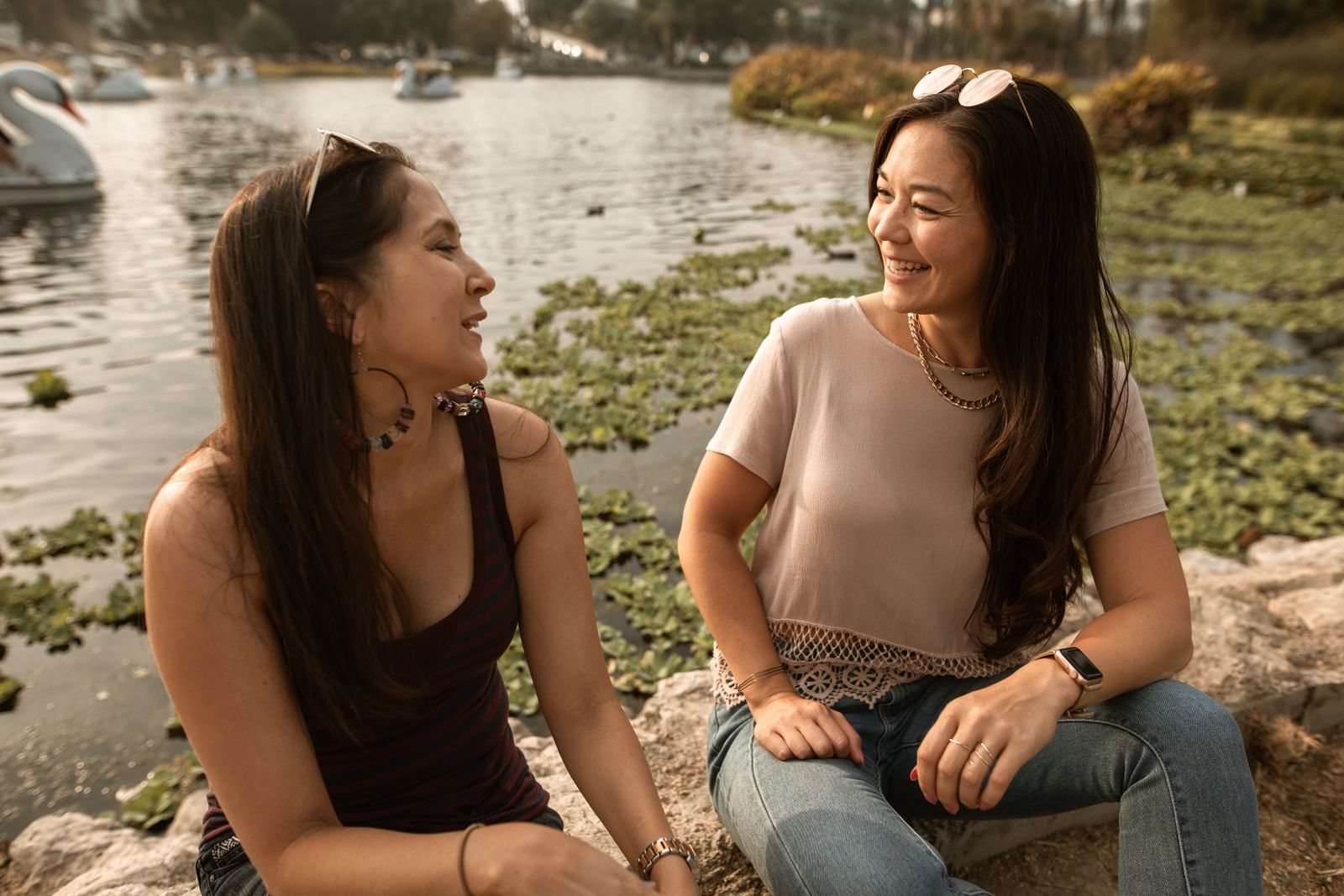 Two women sitting and smiling by the lake, enjoying the outdoors and friendship.