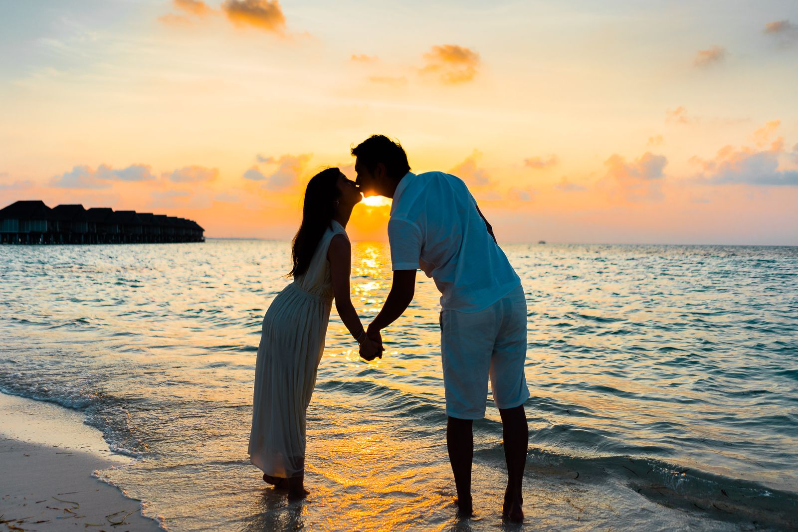 A couple sharing a romantic kiss during sunset on a tropical beach, silhouettes in golden light.