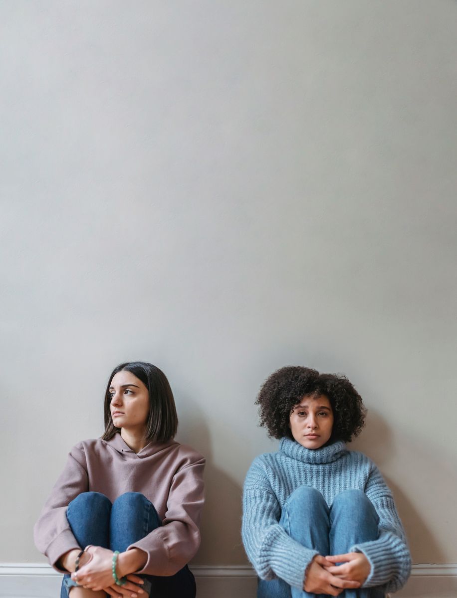 Pensive young multiethnic female friends in casual clothes sitting together while leaning on white wall in light apartment