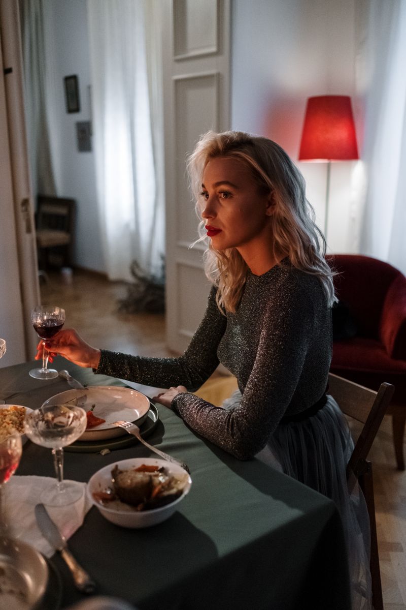 A woman with blonde hair enjoys a cozy dinner indoors, holding a glass of wine.