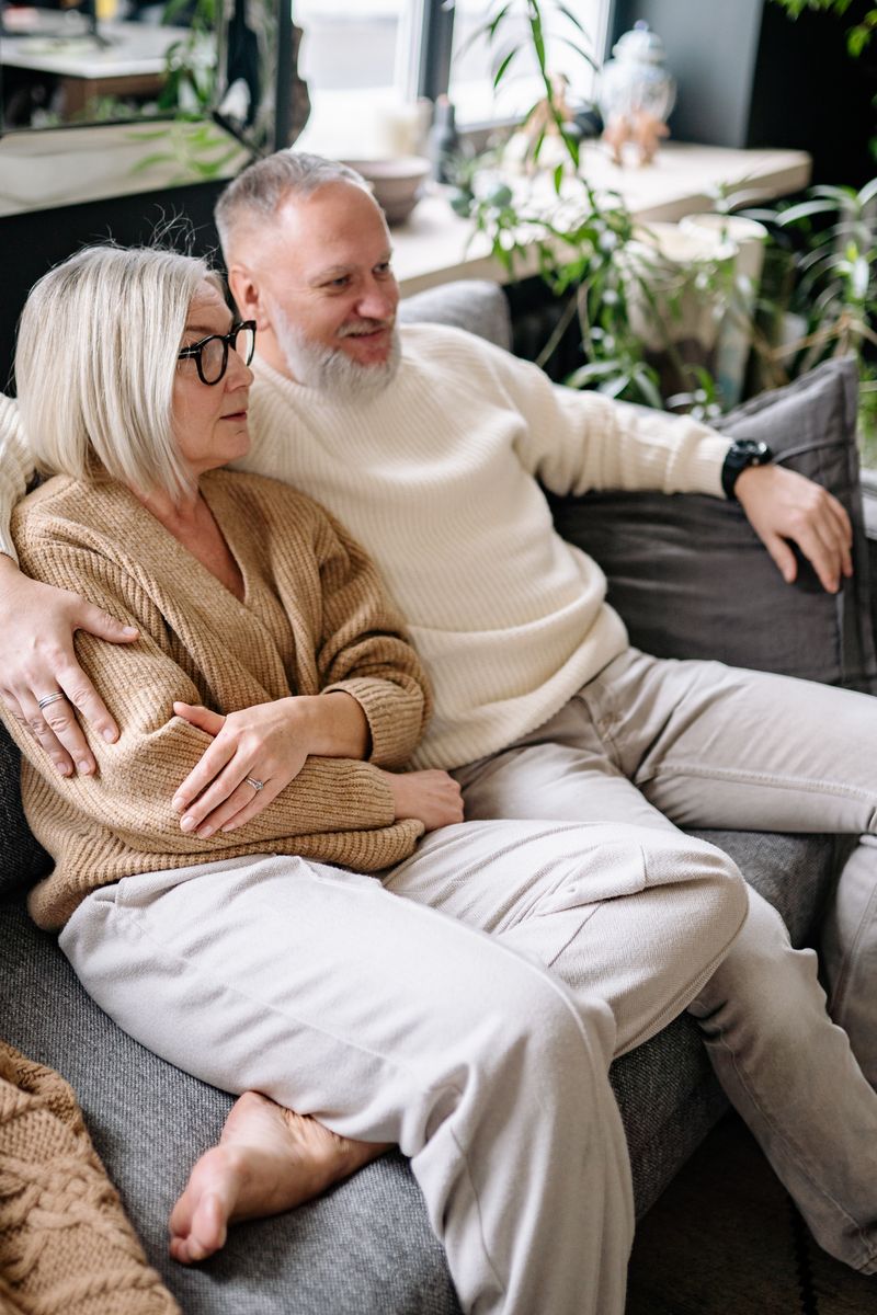 Senior couple sitting on a couch with an arm around each other, enjoying a relaxed moment indoors.