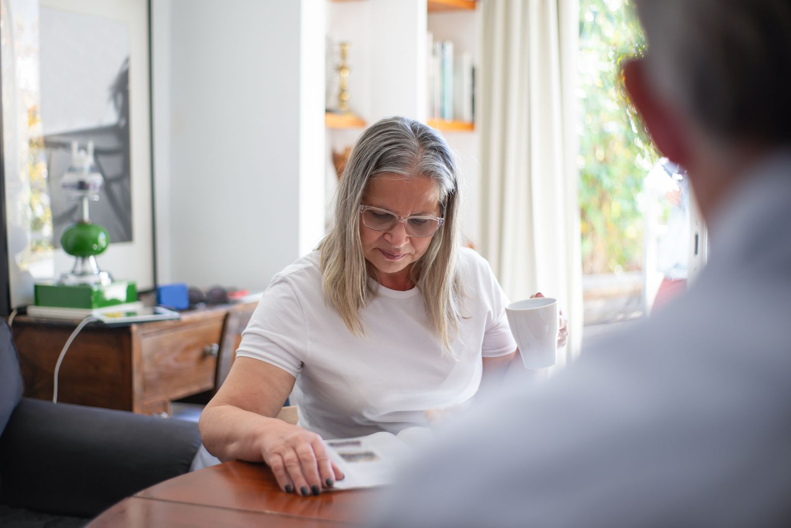 Senior woman wearing eyeglasses reading a magazine with a cup of coffee indoors.