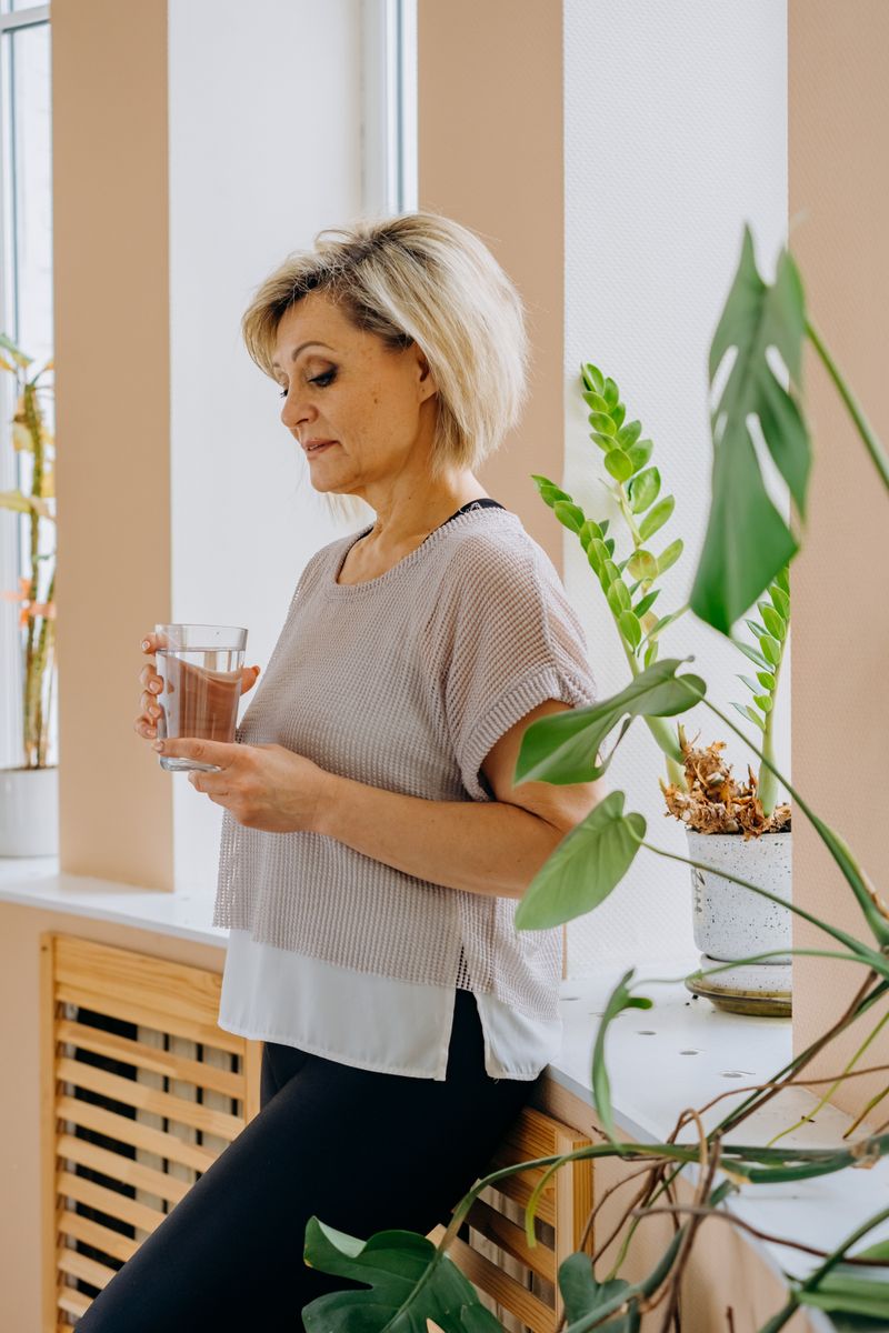 A woman in casual attire holds a glass of water while leaning against a windowsill indoors.