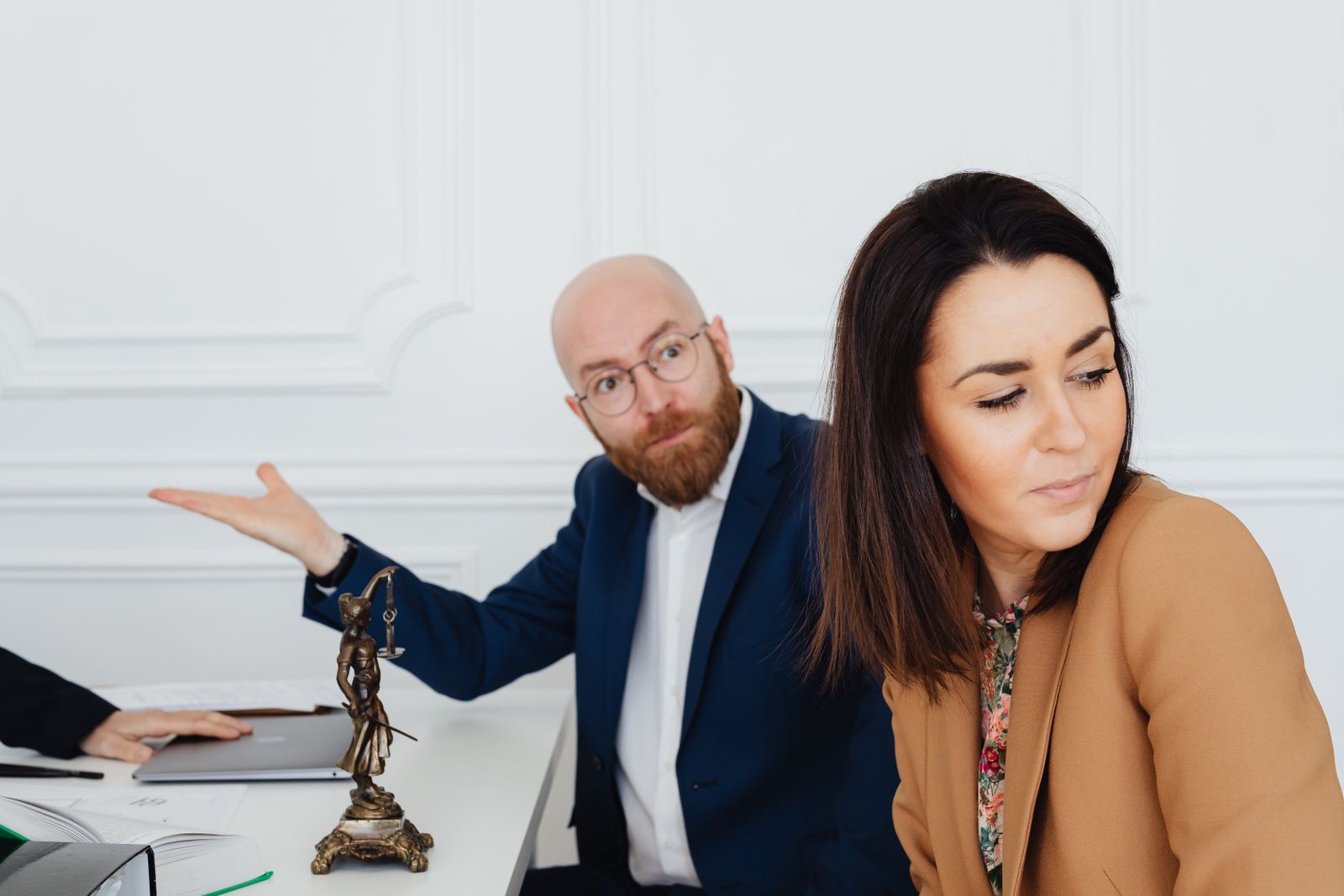 Two professionals in tense discussion beside Lady Justice statue in an office setting.