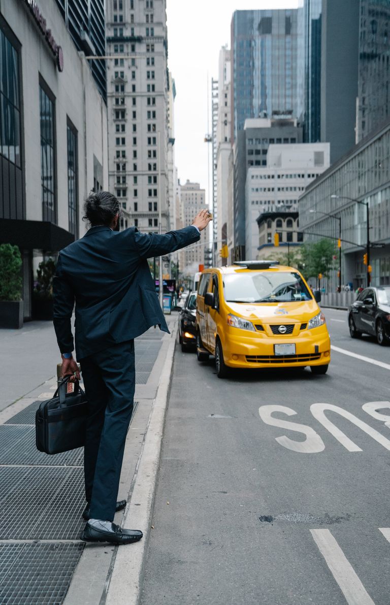A businessman raises his hand to hail a taxi, standing on a bustling city street with skyscrapers in the background.