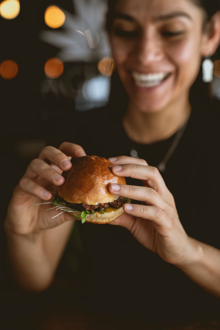 A cheerful woman enjoying a freshly-made hamburger indoors with a blurred background.