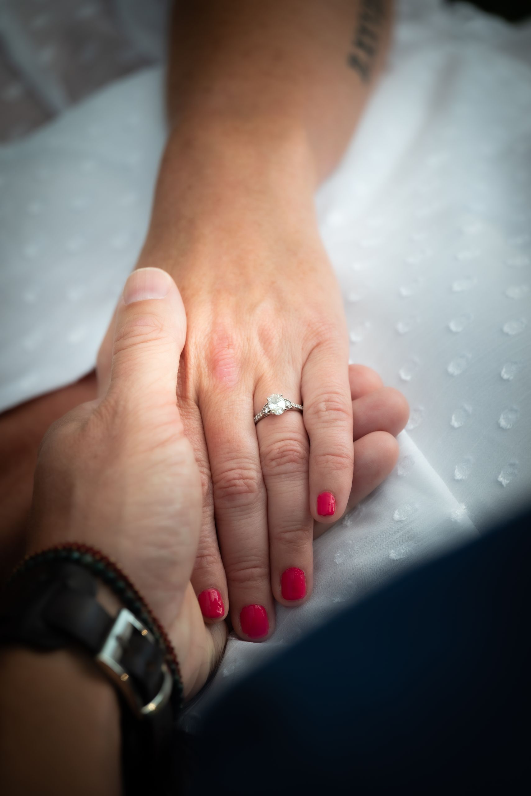 A close-up of two hands holding, highlighting a diamond engagement ring and red manicured nails.