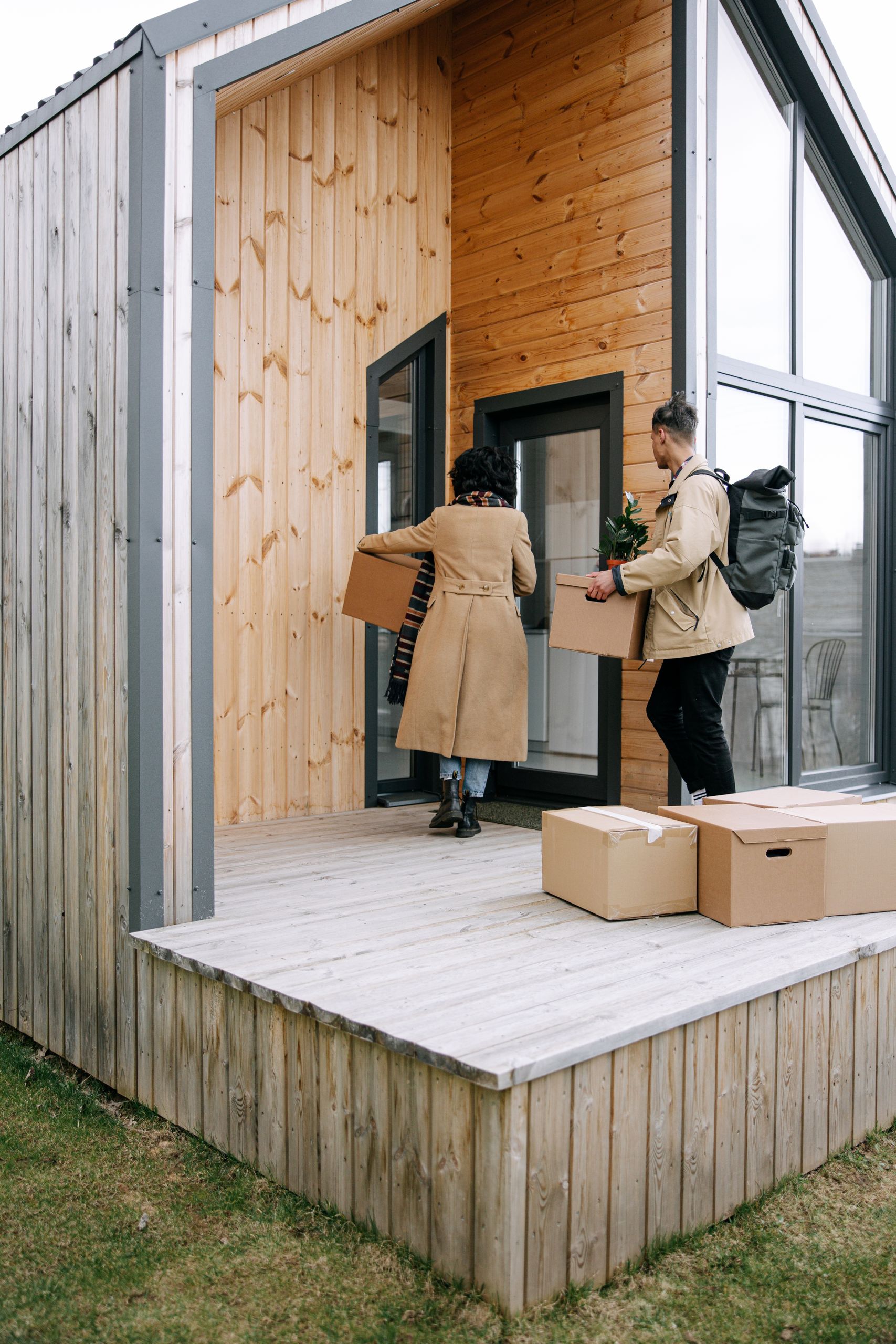 A couple carrying boxes and a plant into their new modern wooden home.