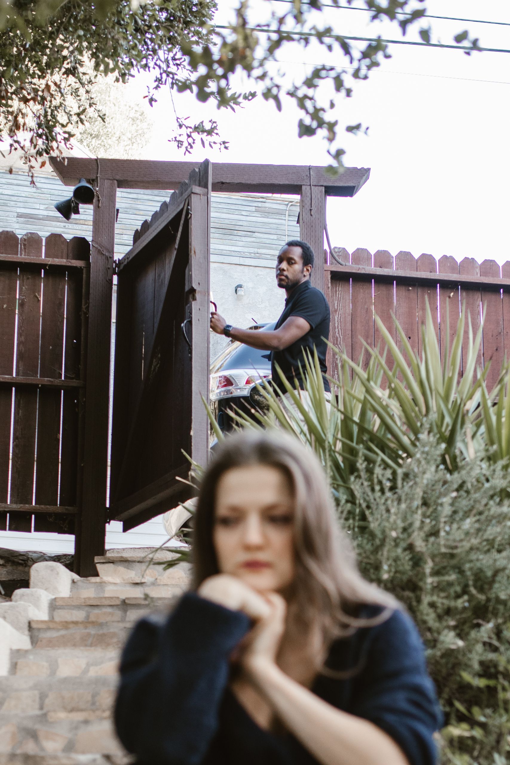 A man and woman face emotional tension and separation outside near a wooden gate.