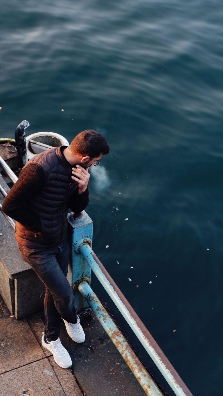 A man leans on a railing by water, smoking in the quiet twilight.