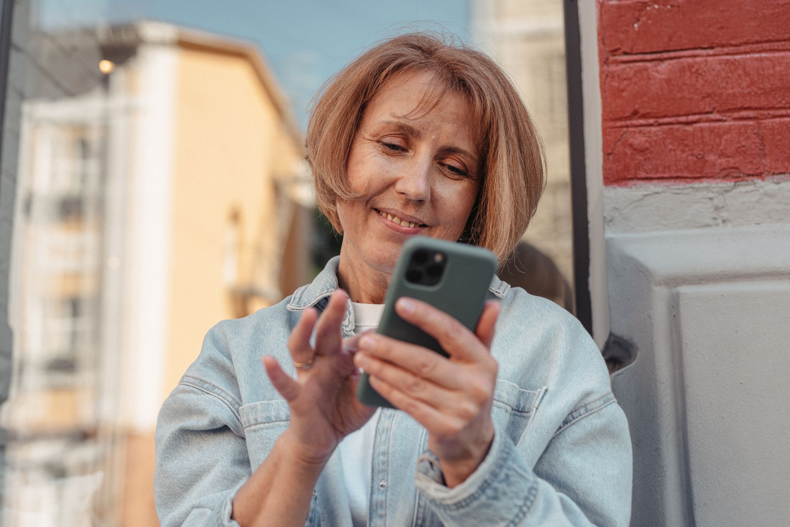 A middle-aged woman in a denim jacket smiles while using her smartphone outside.