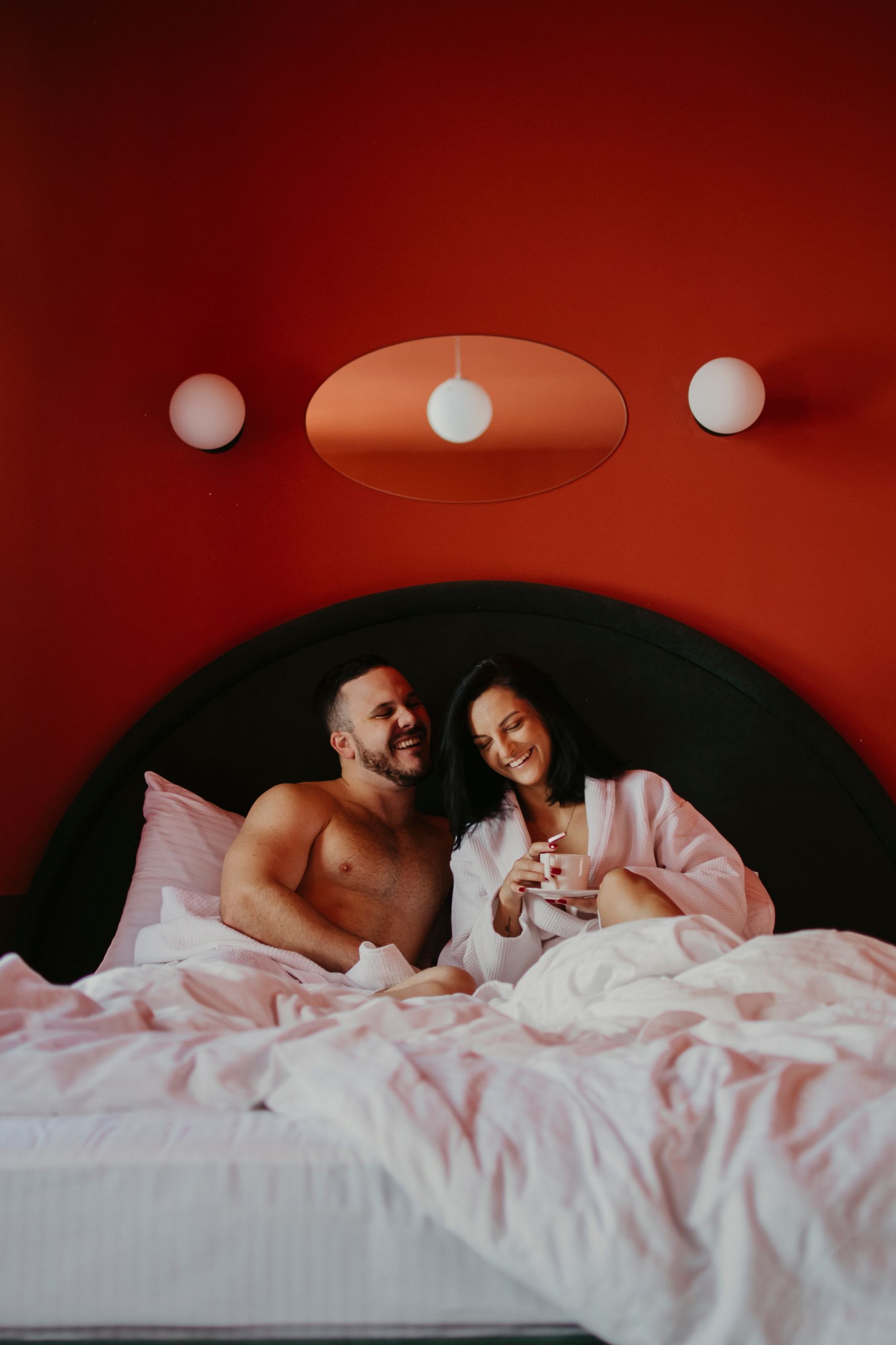 A smiling couple enjoys a cozy morning with coffee in a stylish red bedroom.
