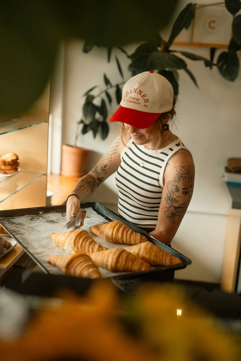 A tattooed female baker carries a tray of freshly baked croissants in a cozy bakery.