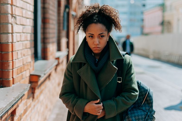 A thoughtful woman in a green coat walks along a sunlit urban sidewalk, lost in contemplation.