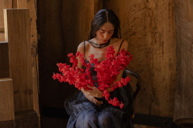 A woman in a dark dress holds vibrant red flowers in a rustic interior, embodying tranquility.