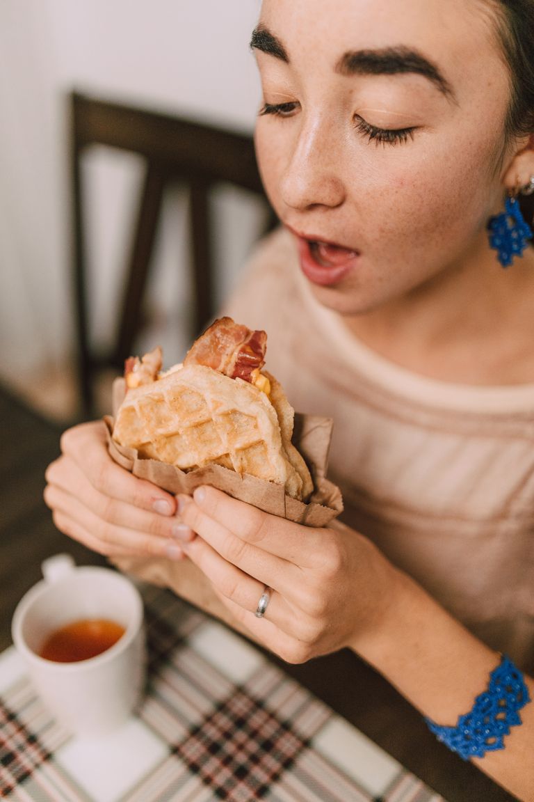 A woman relishes a delicious waffle sandwich with bacon and tea during breakfast.