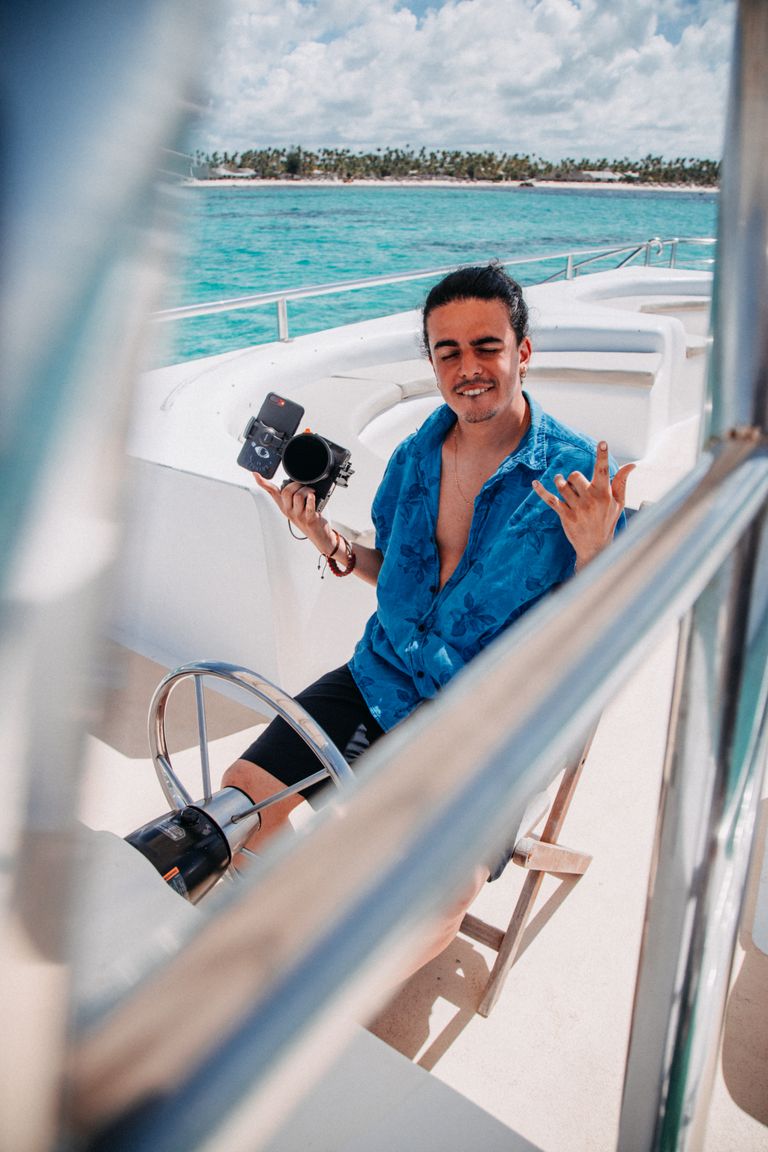 A young man with a camera enjoys a sunny day on a yacht in turquoise waters, capturing moments.