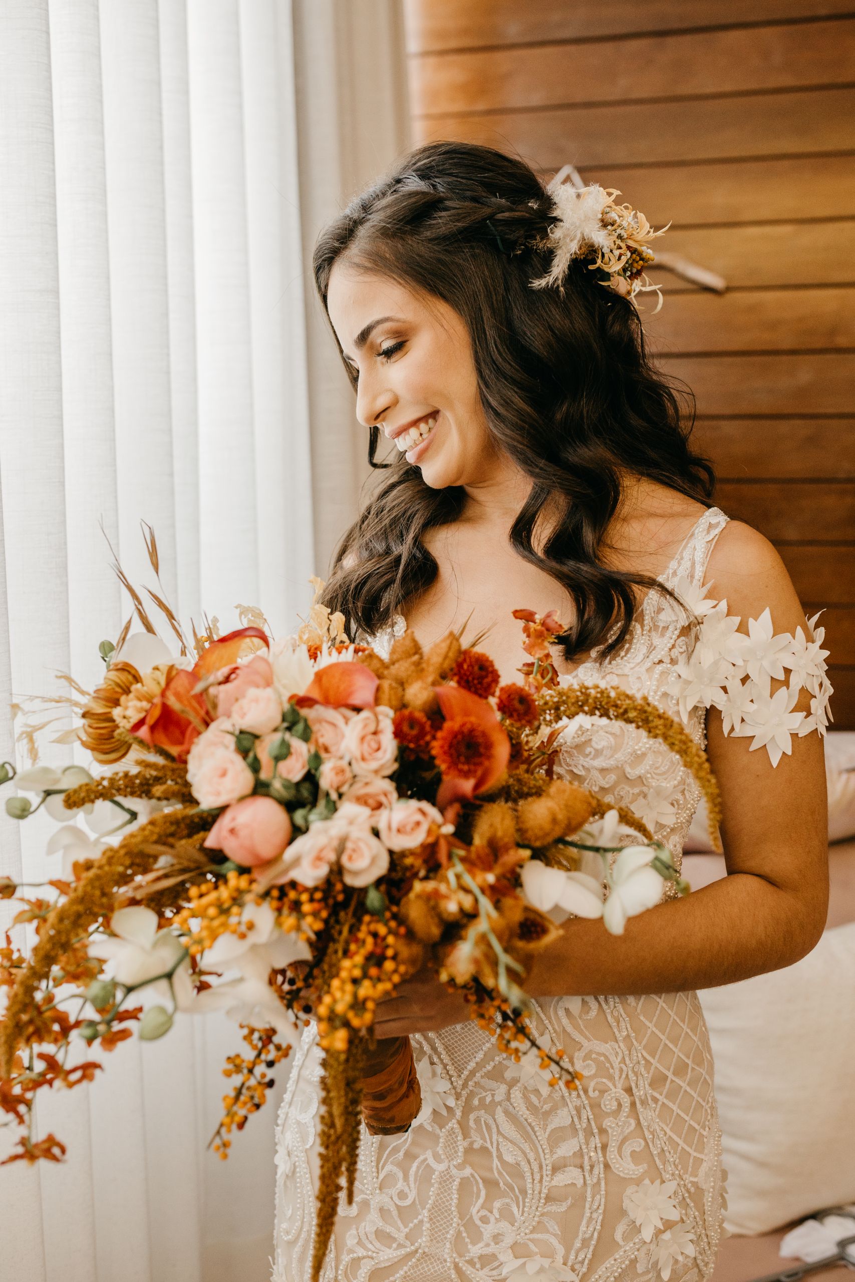 Bride in lace dress holding a stunning autumn-colored floral bouquet, smiling indoors.
