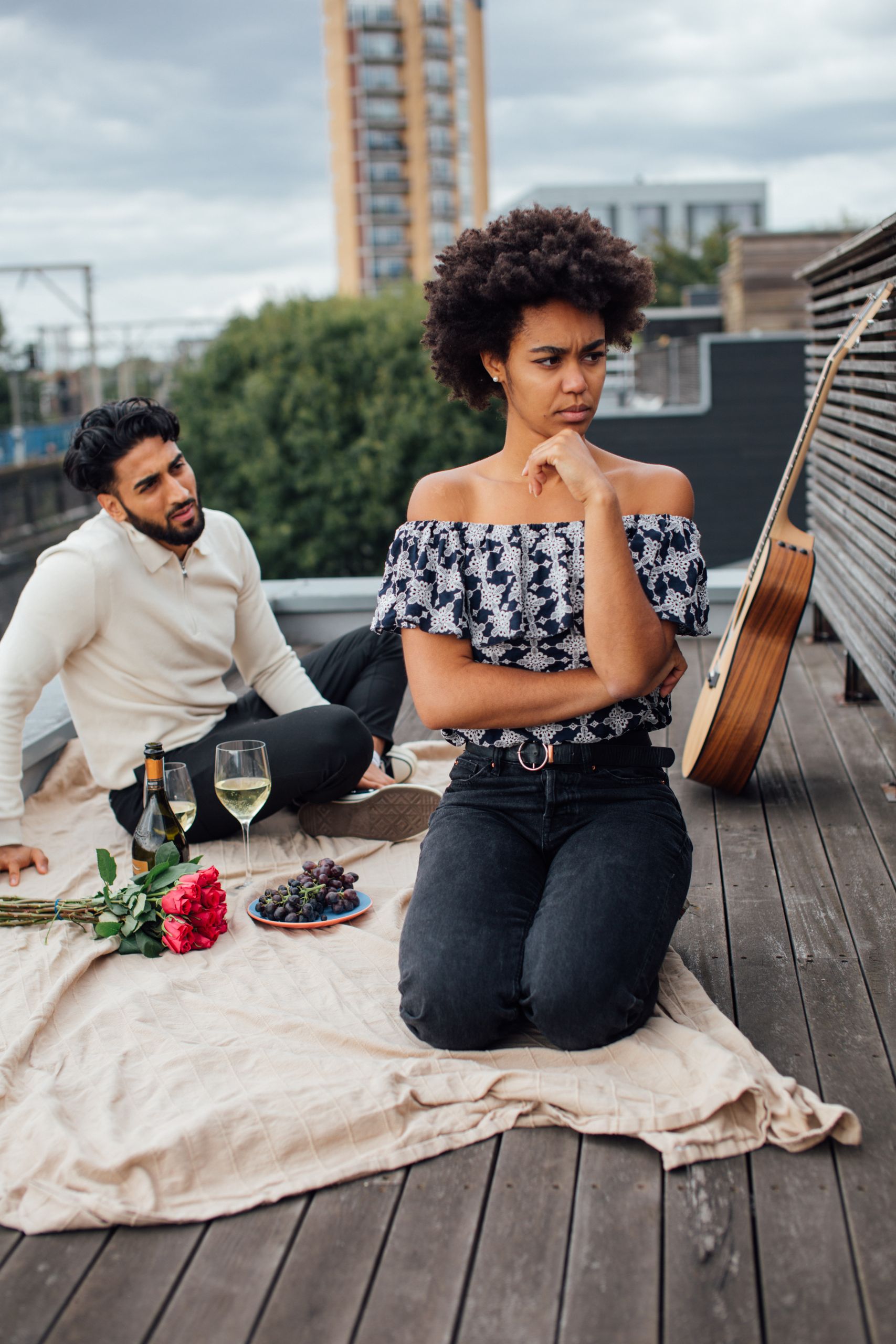 Couple having a romantic picnic on a rooftop, featuring wine, red roses, and an acoustic guitar.