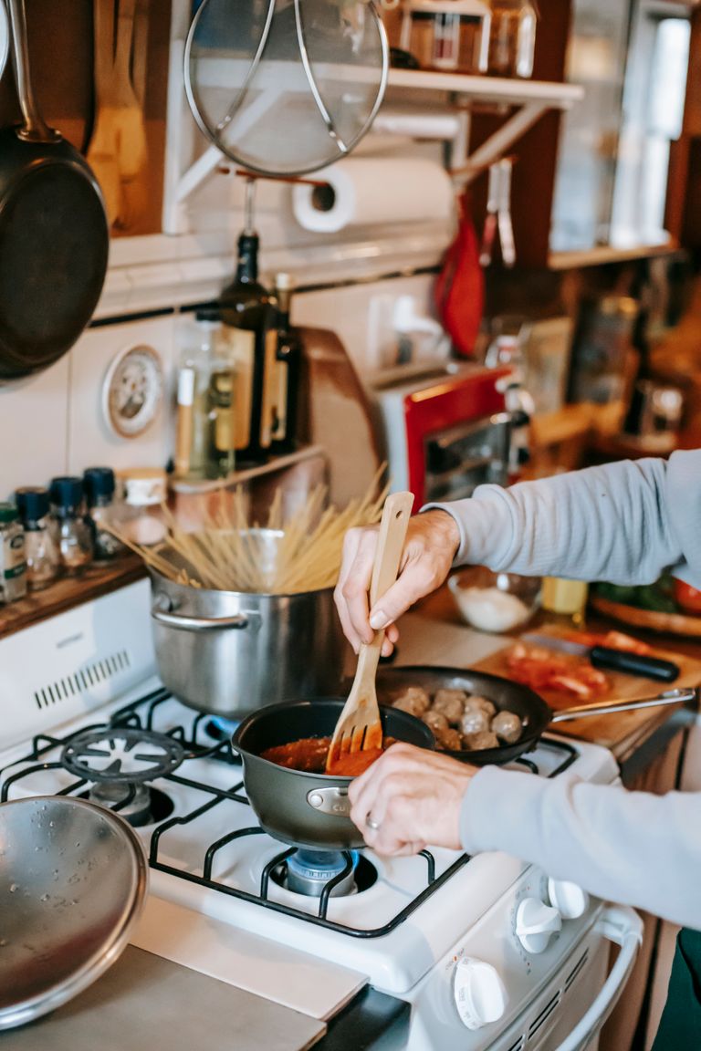 Crop unrecognizable person with spatula and saucepan preparing Bolognese sauce on gas stove against spaghetti at home