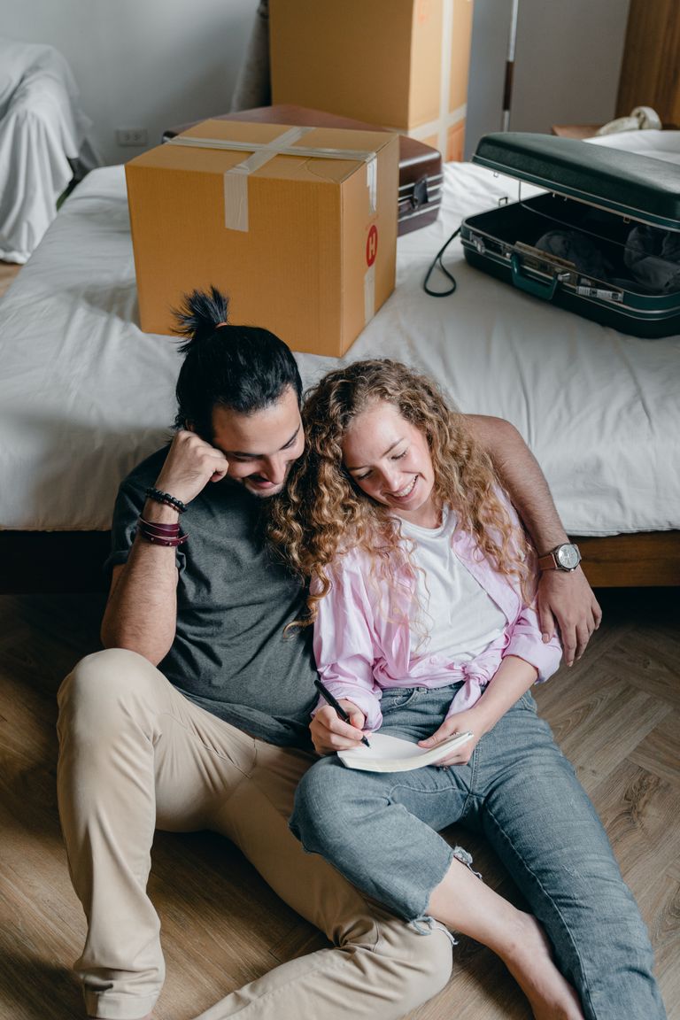 From above of candid couple sitting on floor near bed and writing in notebook while packing things